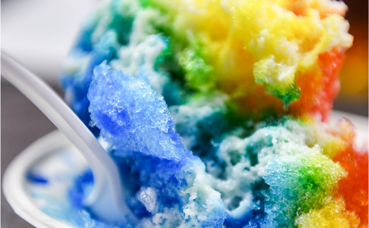 A close up of a spoon dipping into a rainbow colored serving of Hawaiian shave ice.