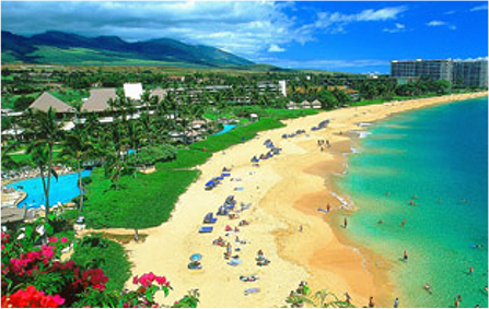 An overhead view of a populated beach in Hawaii. The composition shows the beach is backed by a resort and green mountains.