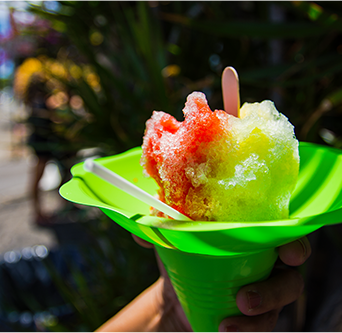A hand holds a serving of shaved ice in the shade. Two flavors have been combined and sit in a plastic cone.
